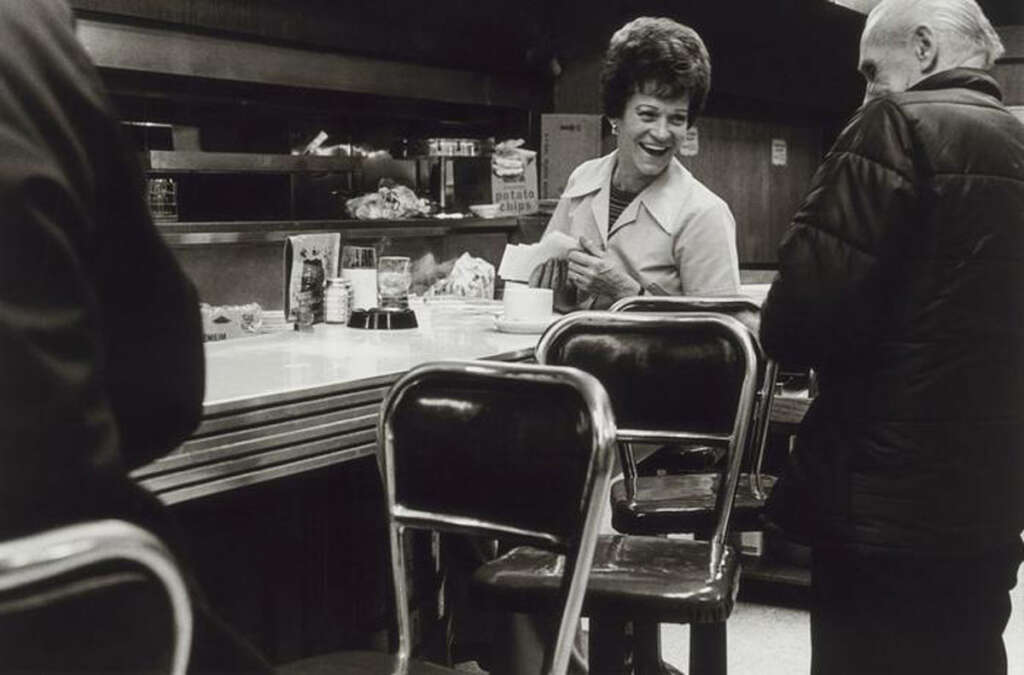 A waitress with short hair smiles while serving a man in a diner. The counter is lined with empty stools and there are various condiments and items on the counter. The man wears a jacket and stands facing the waitress. Black and white photo.