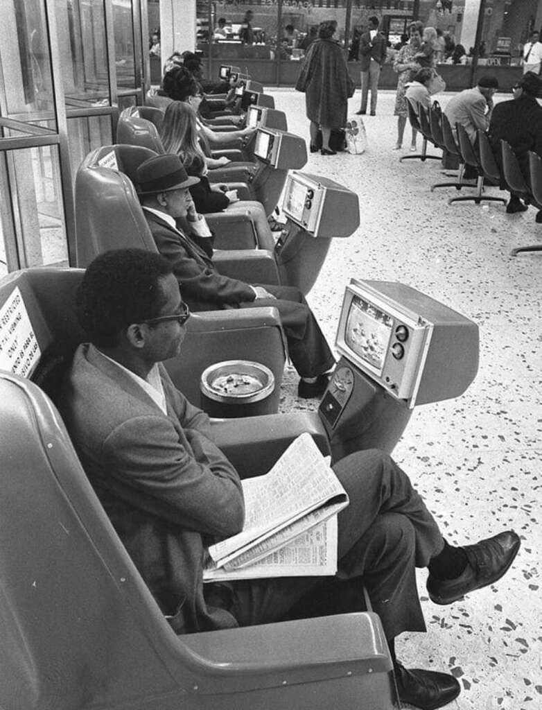 Black and white photo of people seated in retro-style chairs equipped with small TV screens. Some hold newspapers, while others watch TV. The setting appears to be a public space, like an airport lounge or waiting area.