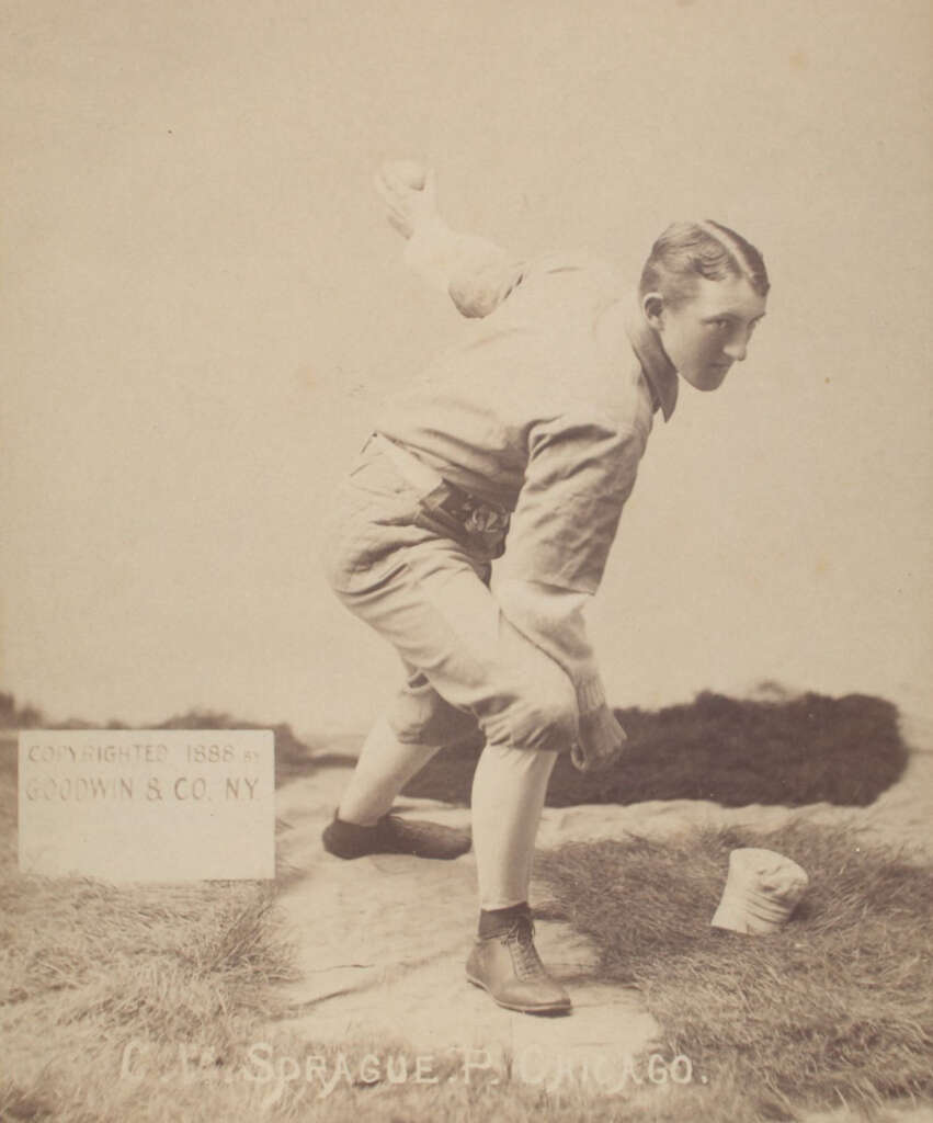 Sepia-toned photo of a vintage baseball player in mid-pitch, wearing a uniform with high socks and a cap. A sign beside him reads "Copyrighted 1888 by Goodwin & Co. N.Y." The player is poised on a grassy area.