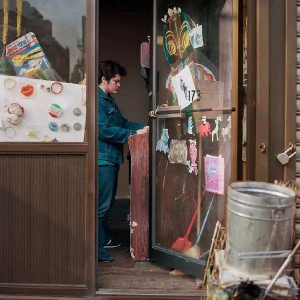 A person stands inside a doorway, partially visible through a glass pane decorated with various stickers and drawings. They are wearing a denim jacket and looking down. A metal container is seen in the foreground outside the door.