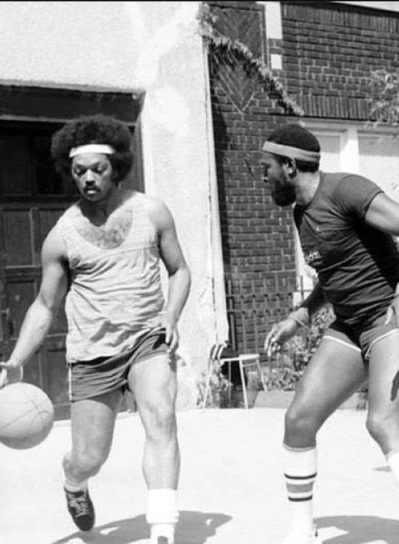 Two men playing basketball outdoors in vintage attire. One dribbles the ball while the other defends. Both wear headbands and sportswear. The setting includes a building in the background. The image is in black and white.