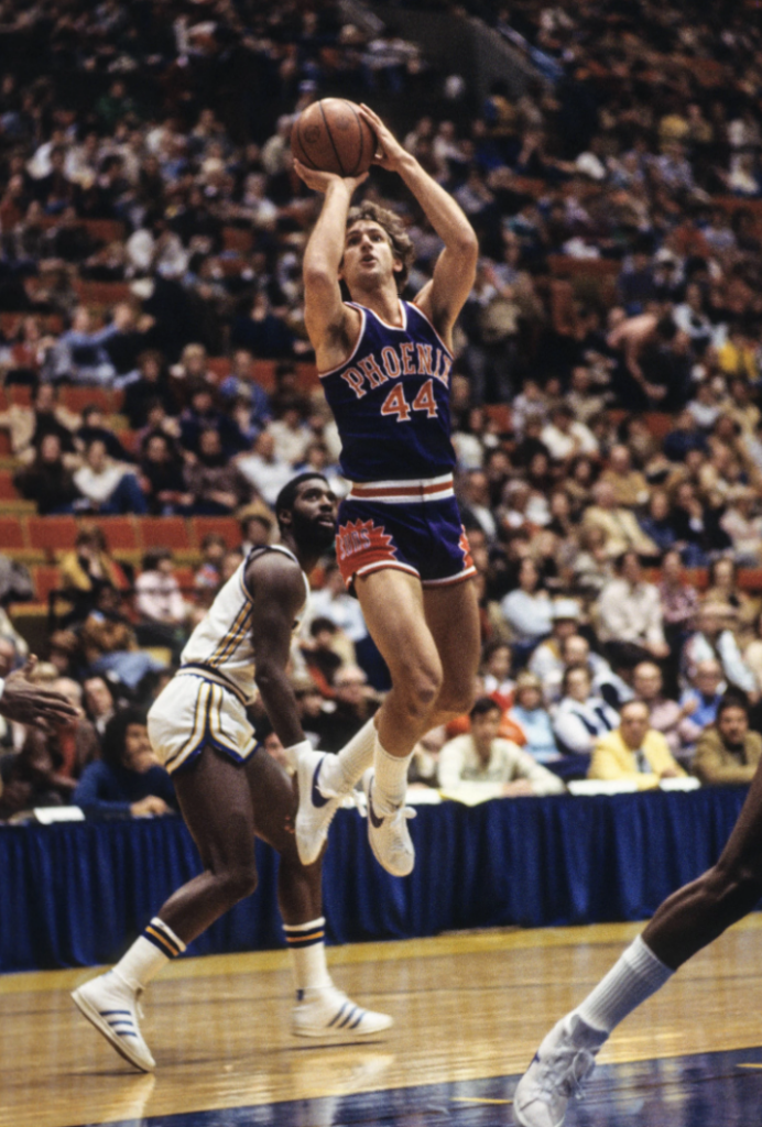 A basketball player in a Phoenix jersey, number 44, jumps to take a shot during a game. An opposing player in a white uniform attempts to defend. The crowd watches from the stands in a packed arena.