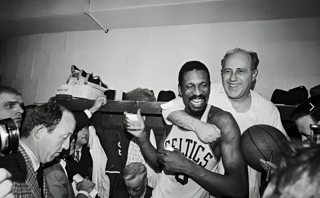 A joyful basketball player in a "Celtics" jersey is celebrated in a crowded locker room. He holds a drink and smiles broadly while being embraced by a man holding a basketball. Cameras and people surround them, capturing the moment.