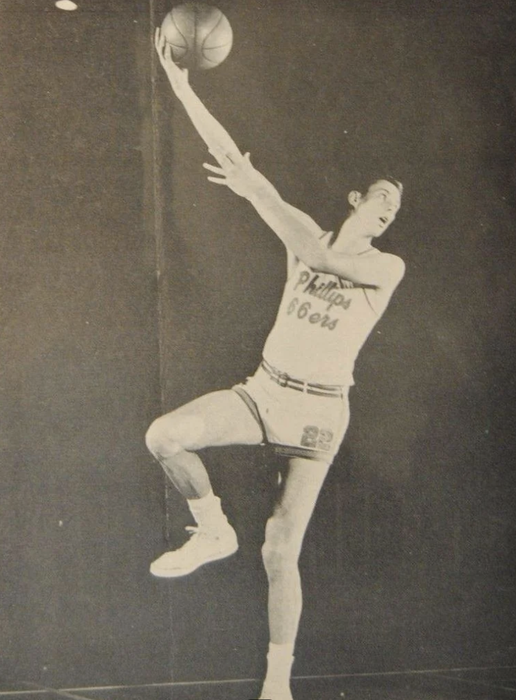 A basketball player in a 76ers uniform jumps and extends his arm to make a one-handed shot. The image is in black and white, capturing a dynamic moment of athleticism.