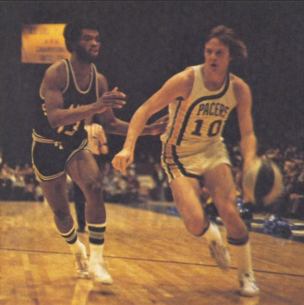 Two basketball players are on a court during a game. One in a white Pacers uniform dribbles the ball while the other in a dark uniform defends. The action takes place in a dimly lit arena with spectators in the background.