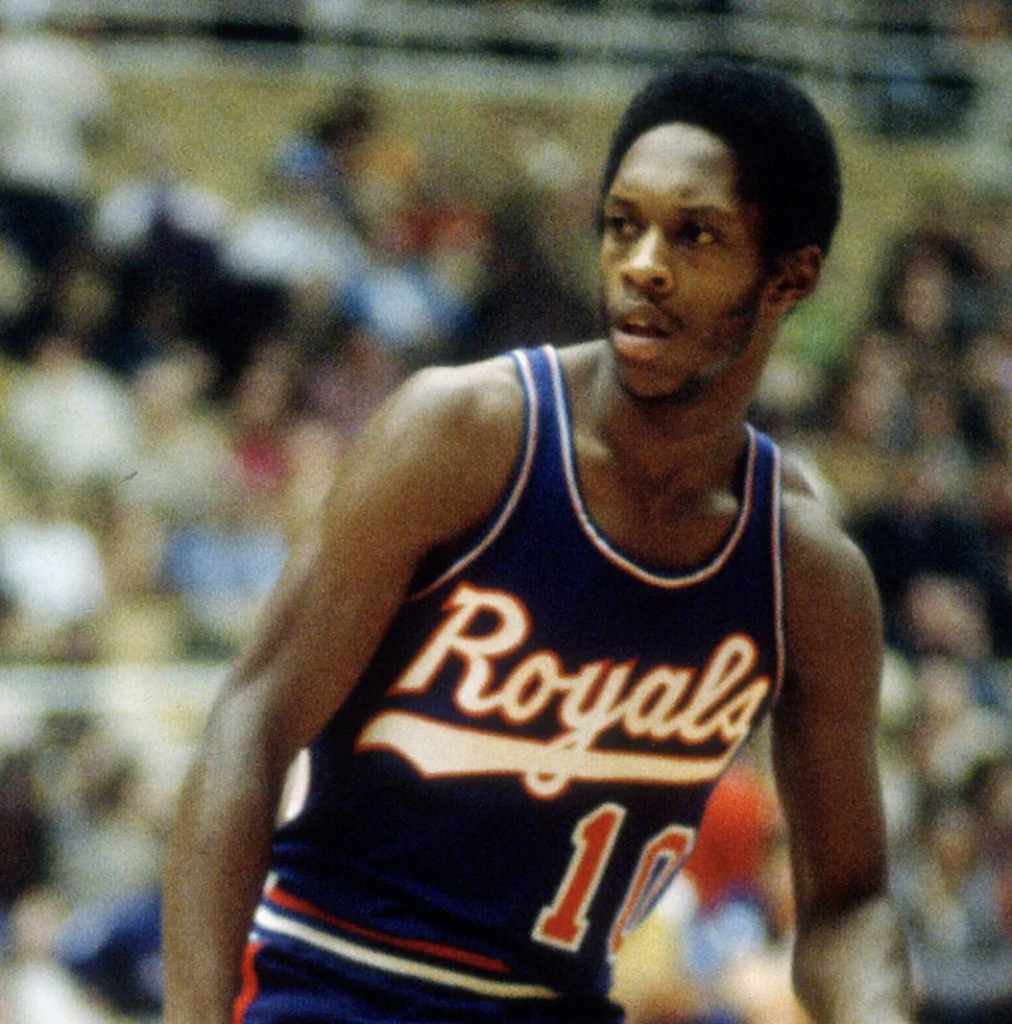 Basketball player wearing a blue Royals jersey with the number 10, standing on a court during a game. The background shows a blurred crowd.