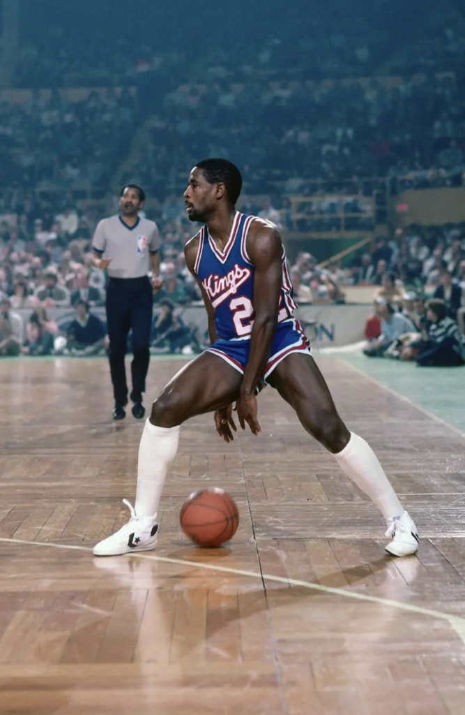 Basketball player in a Kings jersey dribbling on a court, wearing white sneakers, with an official in the background. The audience is watching from the stands.