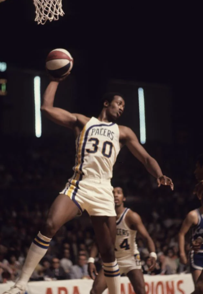 Basketball player wearing a "Pacers" jersey, number 30, holding a basketball with one hand above his head during a game. A teammate is visible in the background on a crowded indoor court.