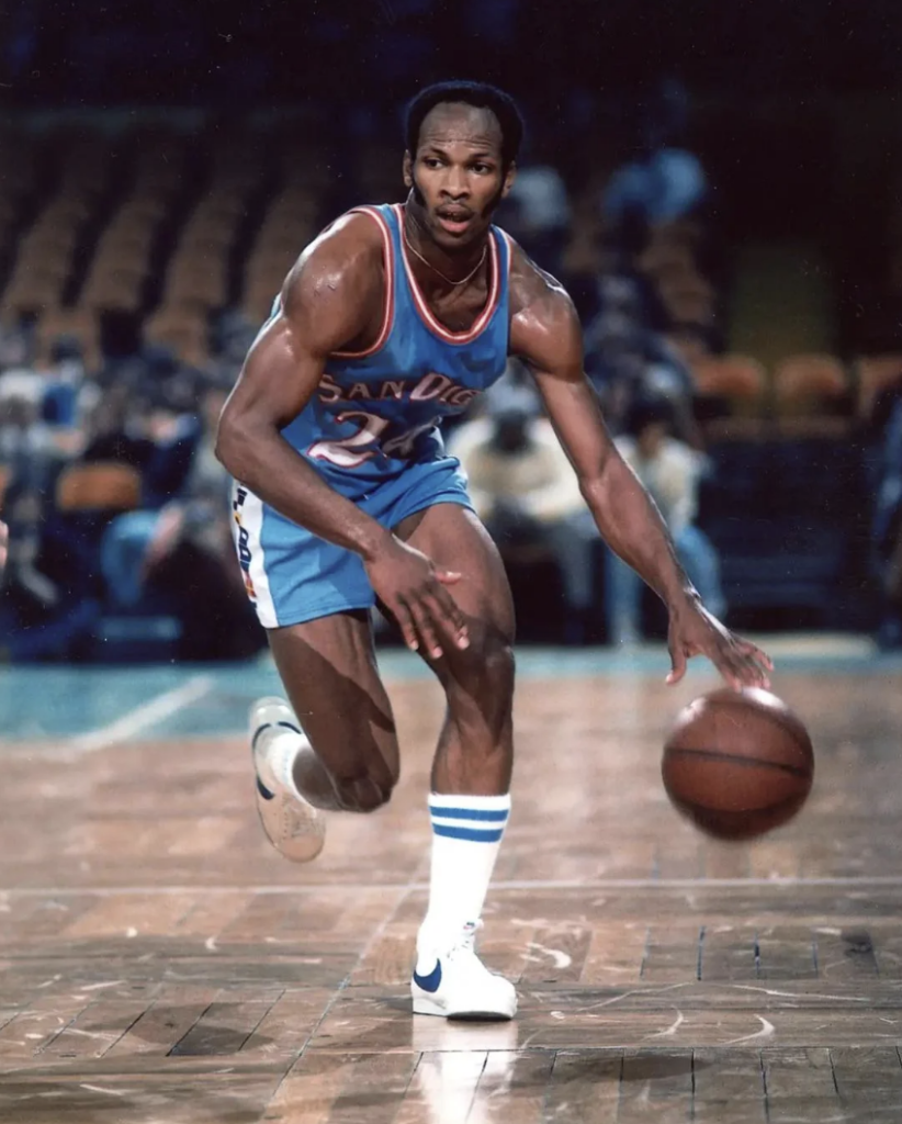 A basketball player in a blue "San Diego" jersey dribbles the ball on a court. The background shows a blurred crowd in the stands. The player is mid-stride, focused, with one hand on the ball.