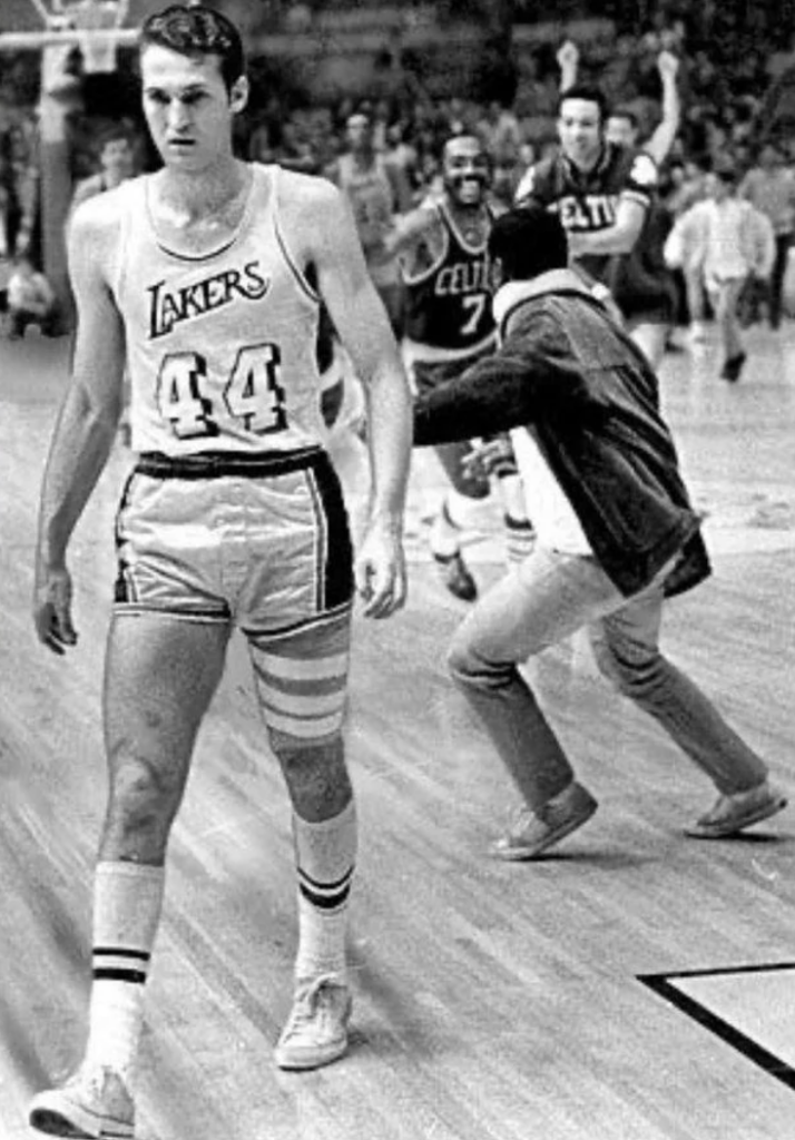 A basketball player in a Lakers jersey with number 44 walks on the court. In the background, people appear to be celebrating, with two individuals in motion, one wearing a Celtics jersey. The scene is from a vintage basketball game.