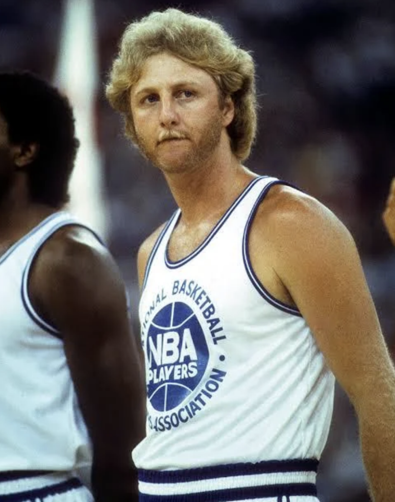 A basketball player with curly blonde hair stands on the court wearing a white jersey with "NBA Players Association" printed on the front. Another player is partially visible beside him.