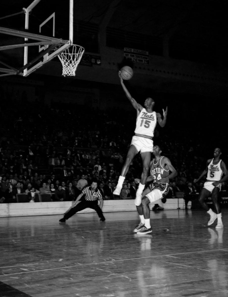 A basketball player, wearing a jersey with the number 15, leaps towards the hoop for a layup as an opponent, wearing number 6, defends. The referee and another player, wearing number 5, watch the action. The scene is set in a crowded indoor stadium.