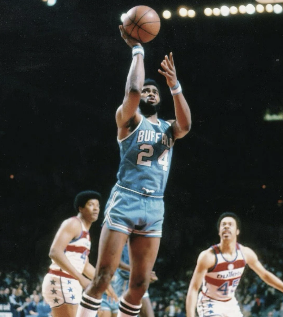 A basketball player in a blue "Buffalo" jersey is jumping to shoot a basketball. Two opposing players in white jerseys watch him. The background shows a crowded arena.