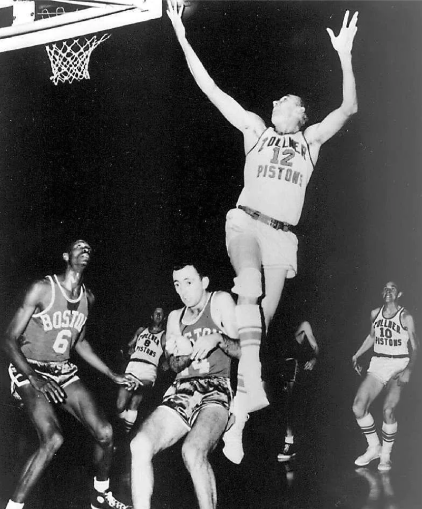 A black and white photo of a basketball game. A player in a Pistons jersey leaps towards the basket. Opponents in Boston jerseys are near the basket, looking up. Other players observe from a distance.