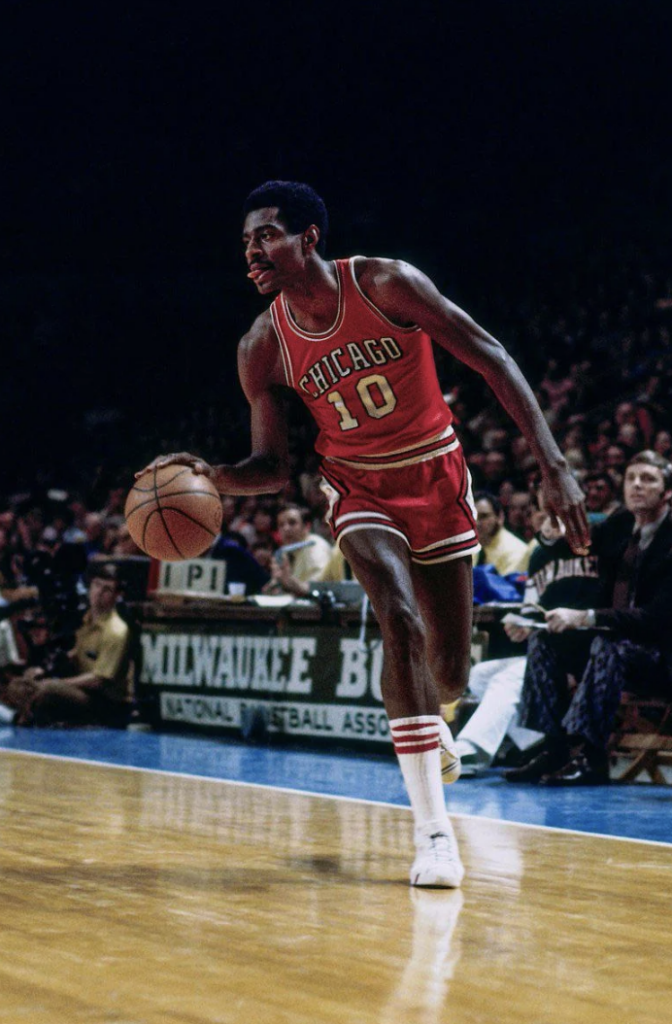 Basketball player dribbling a ball on a court. He wears a red Chicago uniform with the number 10. Spectators are visible in the background. A sign reads "Milwaukee Bucks National Basketball Association.