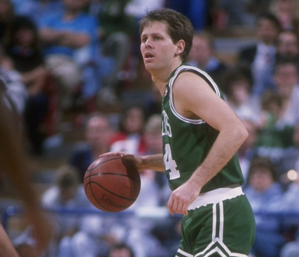 Basketball player in a green and white uniform dribbles the ball on a court. The audience is blurred in the background.