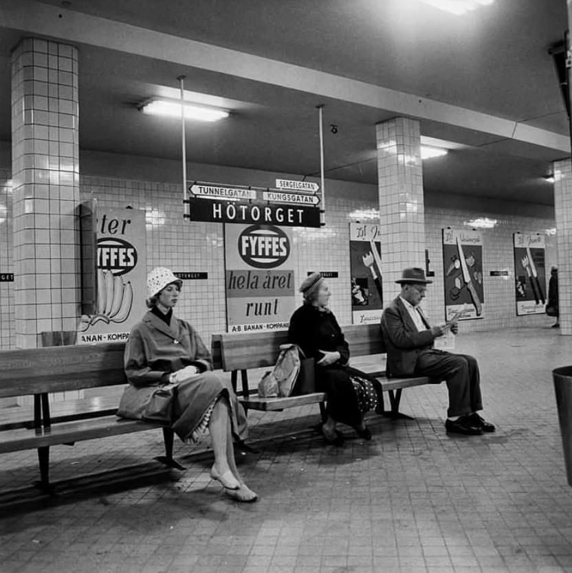 Black and white photo of three people sitting on a bench in a tiled subway station. They appear to be reading newspapers. Behind them, vintage posters and signs are visible on the walls. The atmosphere is calm and quiet.