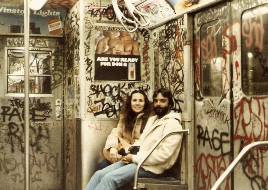 A couple sits on a graffitied subway train bench, smiling. The walls are covered in colorful tags and drawings. A poster on the wall reads, "Are you ready for DK 86?" with graffiti all around. The train door is visible in the background.