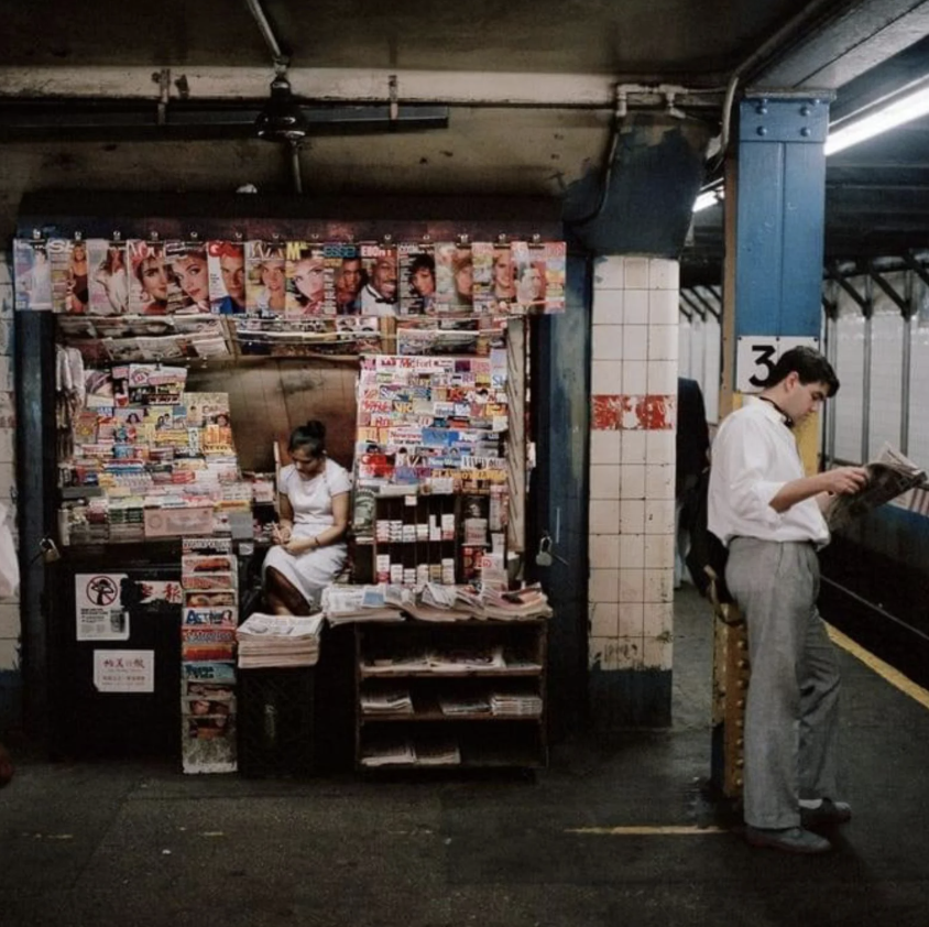 A newsstand in a subway station is packed with magazines and newspapers. A person is sitting inside reading. Another person stands nearby, leaning against a column and reading a newspaper, as they wait for a train.