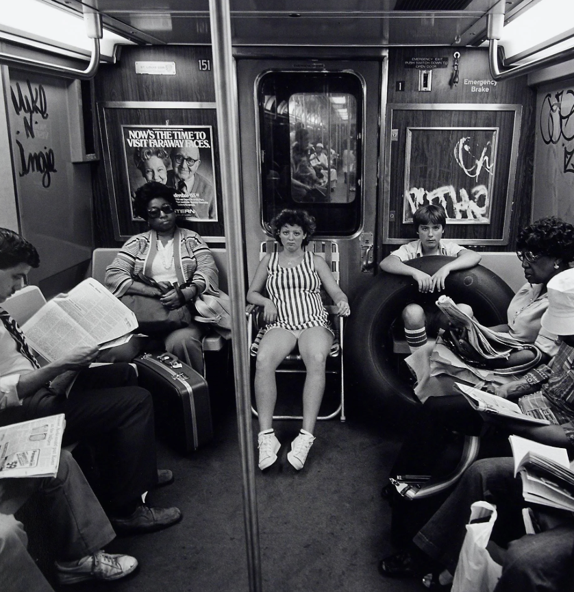 Black and white photo of a subway car with several passengers. A woman in a striped outfit sits on a folding chair in the center. Other people are reading, and a girl with an inner tube sits nearby. Graffiti is seen on the walls.