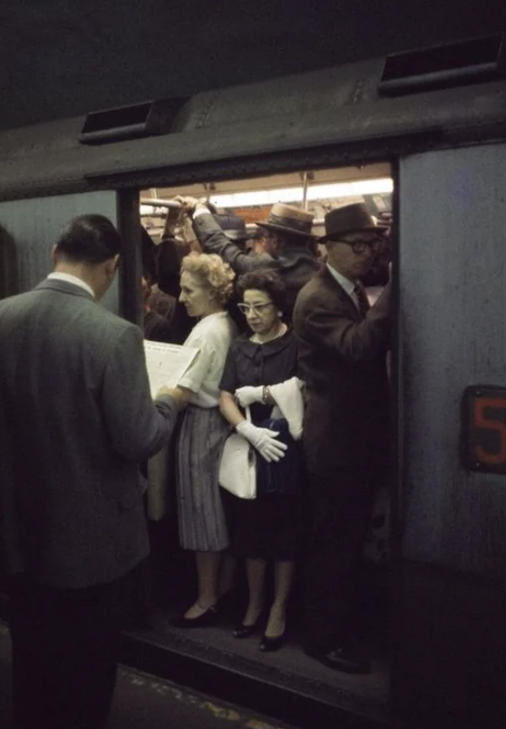 Crowded subway train with passengers standing closely. A man reads a newspaper, and a woman in gloves and an older man with a hat are visible near the door. The train appears full, capturing a typical busy commute.