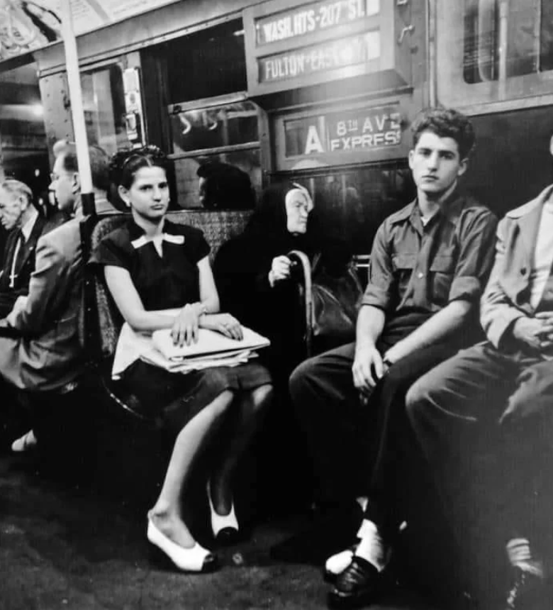 Black and white photo of passengers seated inside a subway train. A young woman in a dress sits with clasped hands, next to an elderly woman with a cane. A young man in a military uniform sits beside them. Advertisements line the train walls.