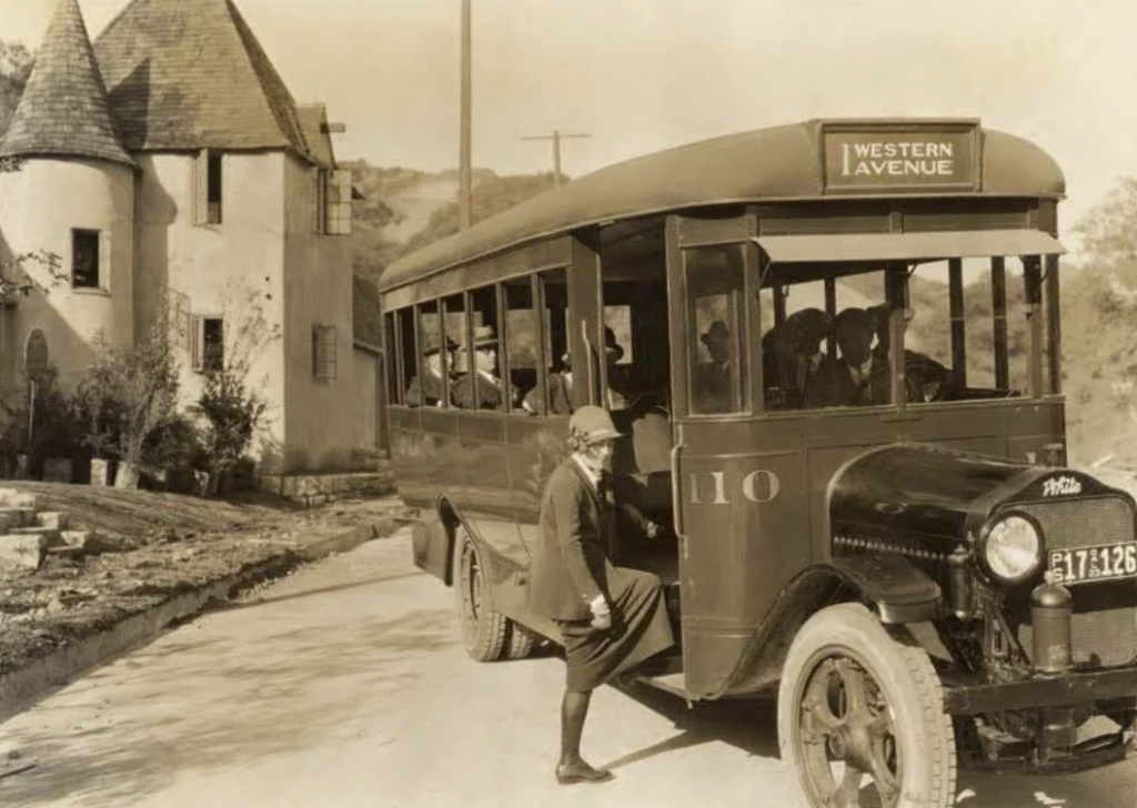 A vintage bus labeled "Western Avenue" is parked on a road beside a castle-like building. The bus is filled with passengers, and a man in a suit and hat is stepping up to board. The scene appears to be from the early 20th century.