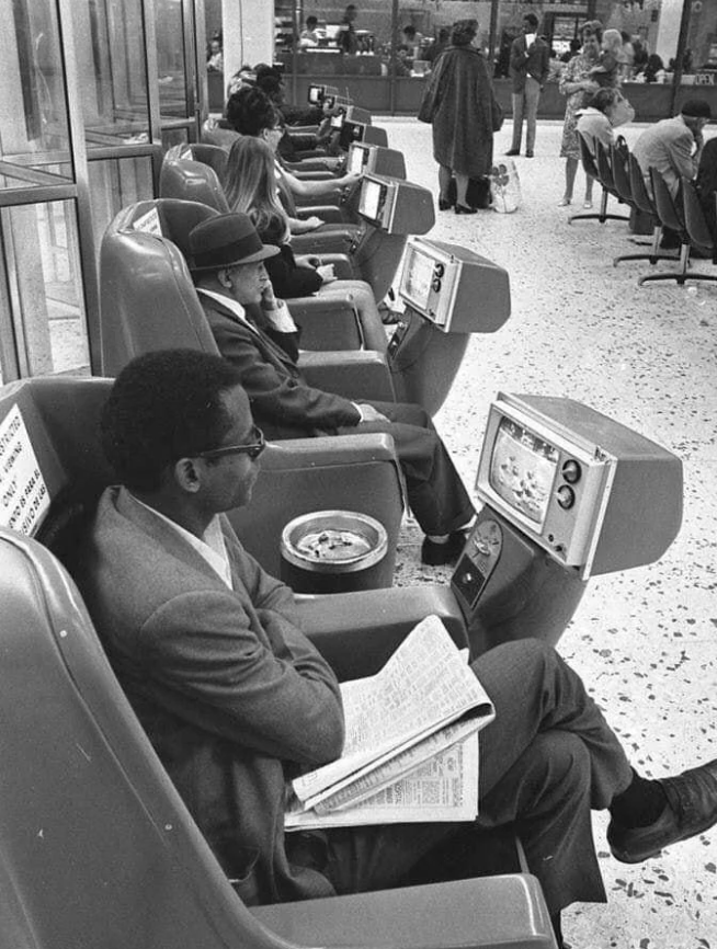 People seated in rows of futuristic chair pods with individual televisions, some reading newspapers. They appear in a public setting with large windows, possibly a transportation hub. The decor is retro, suggesting a scene from the past.