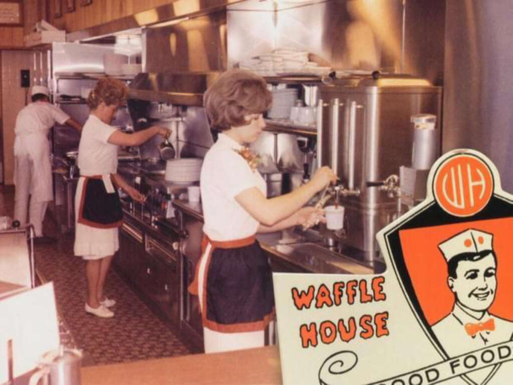 A vintage diner scene with two women in uniforms working behind a counter, preparing food and drinks. The decor is retro, with kitchen equipment and shelves of dishes visible. A Waffle House sign with an illustration of a smiling chef is in the foreground.