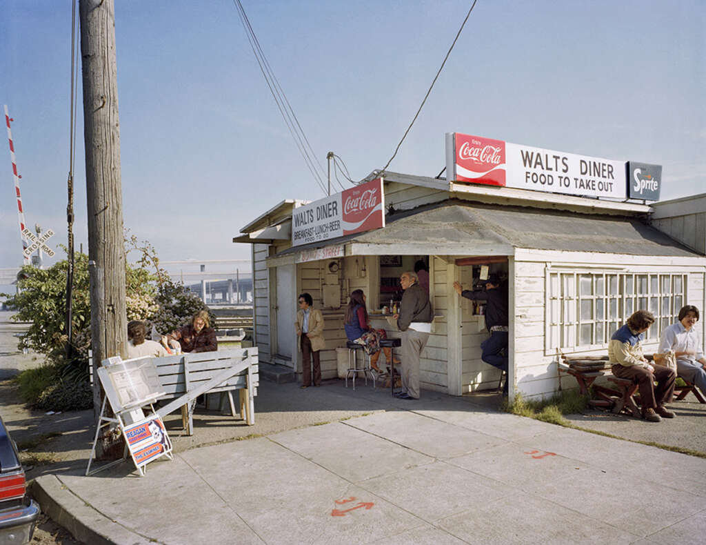 A small, rustic diner with a sign reading "WALTS DINER FOOD TO TAKE OUT" is on a street corner. Several people are sitting and standing outside. A few signs advertise Coca-Cola and Sprite. A utility pole and the road are in the foreground.