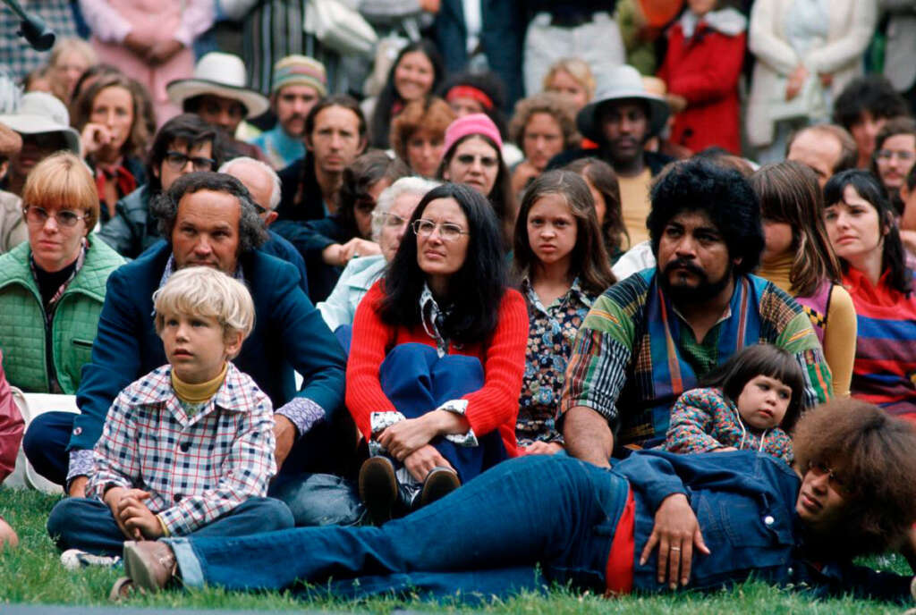 A diverse group of people sit closely together on the grass at an outdoor event. They appear engaged, with various expressions and styles of clothing. There are adults and children present, creating a warm, communal atmosphere.