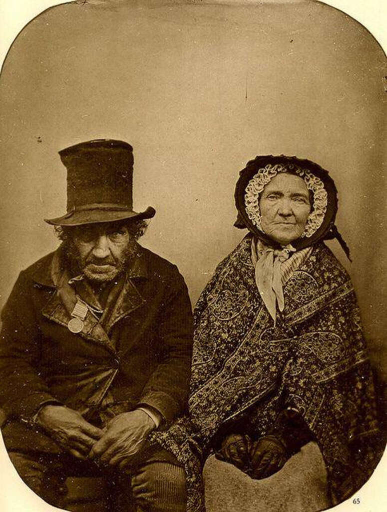 Vintage sepia-toned photograph of an elderly couple. The man wears a tall hat and coat, looking serious, with a medal around his neck. The woman is draped in a patterned shawl and bonnet, sitting beside him, appearing calm.