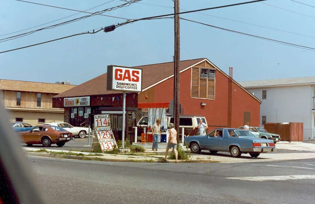 A small gas station with a red building offers gas, sandwiches, deli, and coffee. A white van and several cars are parked nearby. The gas prices are shown on a sign. People are walking and standing around, with buildings in the background.