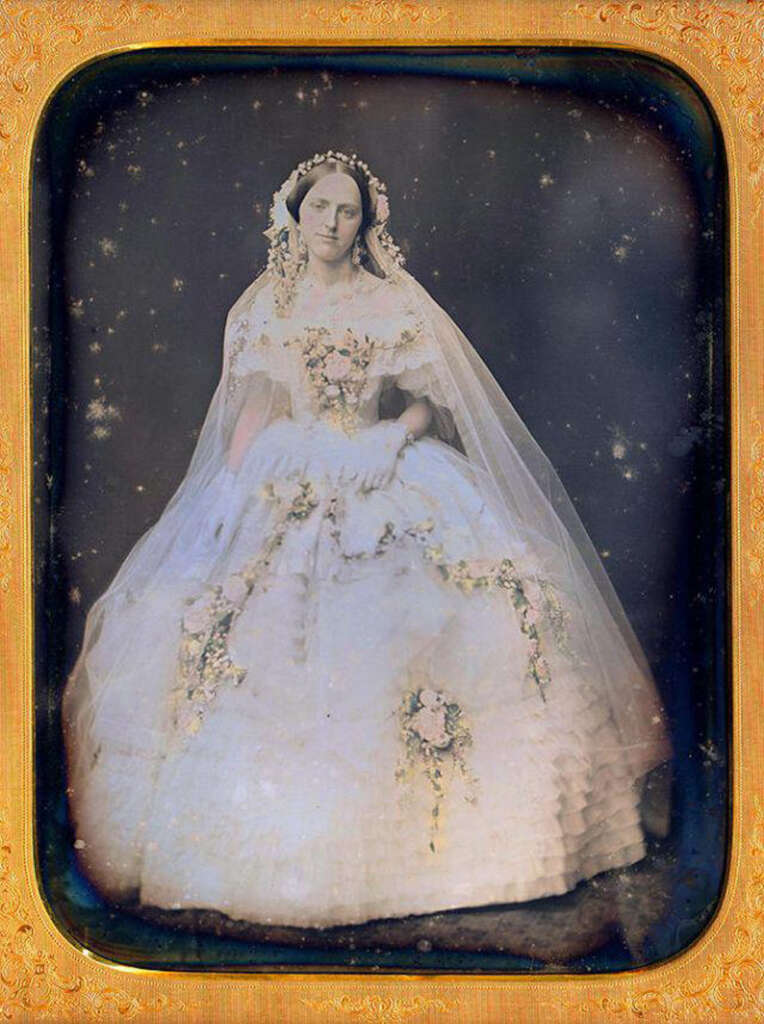 A vintage photograph of a woman in an ornate dress with floral embellishments. She wears a veil and floral headpiece, sitting posed with hands resting on her lap. The image has a decorative gold frame typical of daguerreotypes.