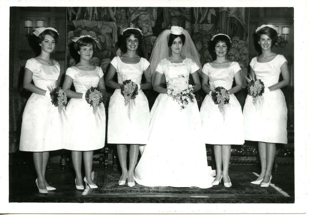 A bride stands smiling in the center, wearing a long white gown and veil, holding a bouquet. She is flanked by five smiling bridesmaids in matching short dresses and holding bouquets. They are posing indoors against an elegant backdrop.