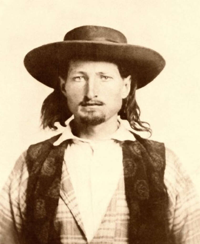 Sepia-toned vintage portrait of a man with long hair wearing a wide-brimmed hat, a patterned vest, and a collared shirt. He has a mustache and goatee, gazing directly at the camera.