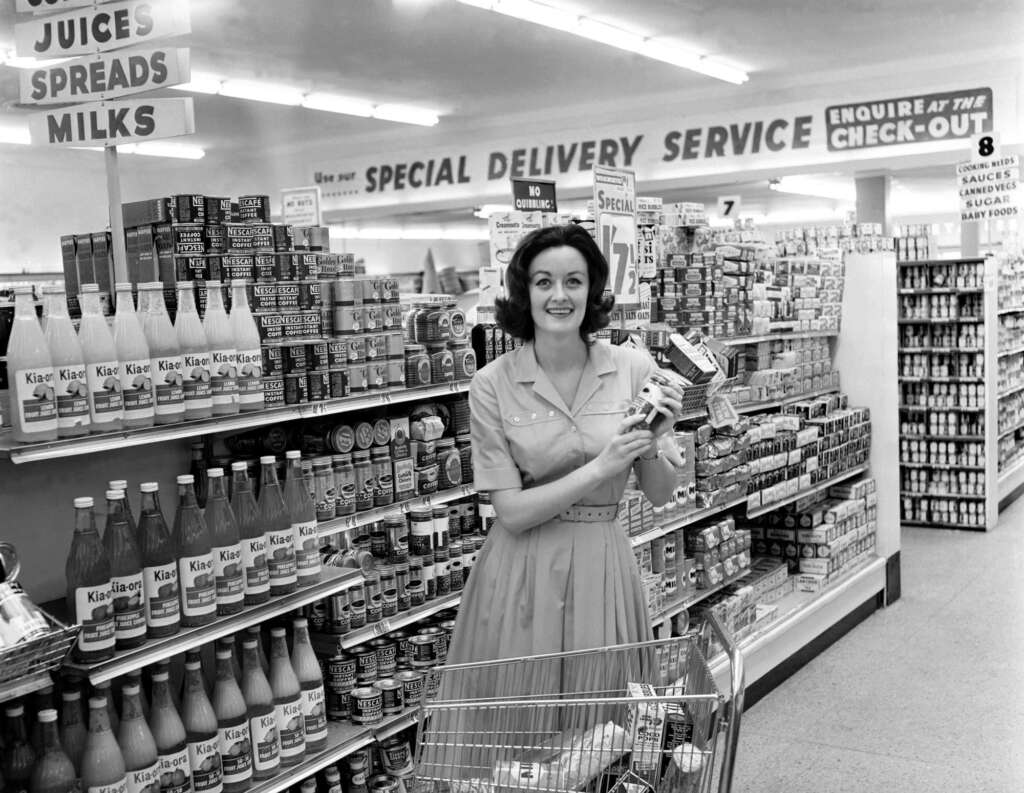 A woman in a dress smiles while holding packaged items in a grocery store. She stands near a cart in an aisle displaying various products like juices and sauces. Signs above mention "Special Delivery Service" and "Juices, Spreads, Milks.