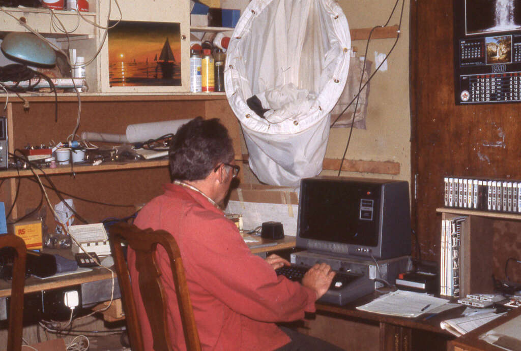 A person in a red jacket sits at a cluttered desk using a vintage computer. Shelves are filled with various items, including a painting of a sunset with sailboats. A white fabric object hangs on the wall, and a calendar is visible nearby.