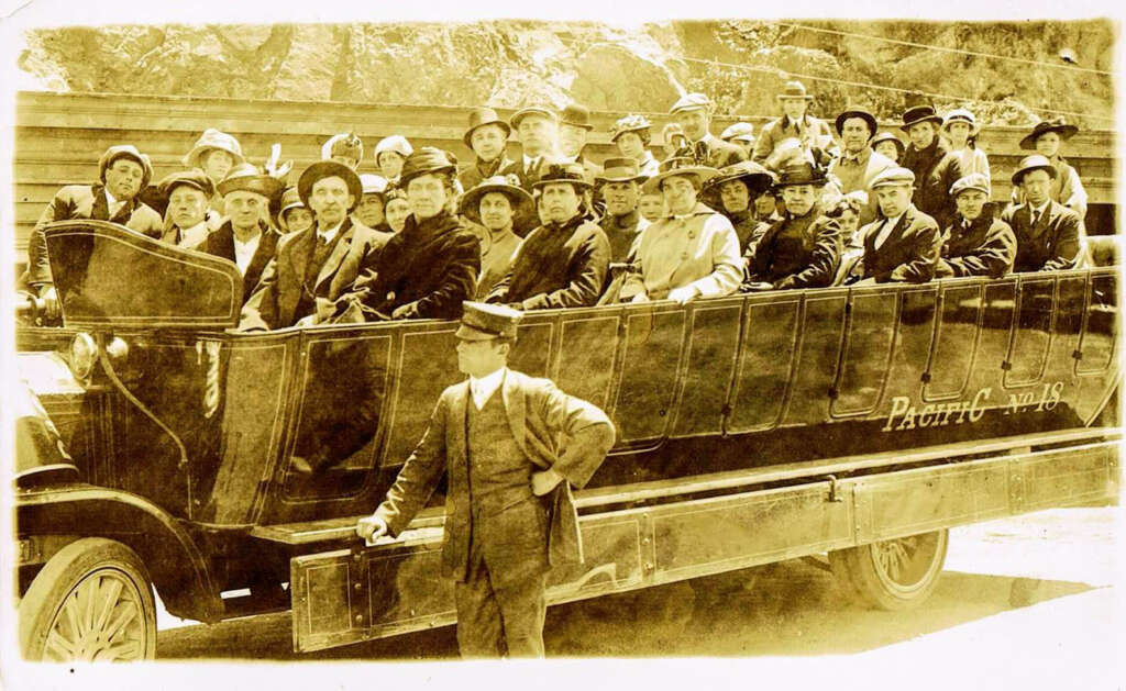 A sepia-toned vintage photo of a large open-sided vehicle filled with men and women wearing hats and suits. The vehicle is labeled "Pacific No. 48." A man in a suit and hat stands in front of the vehicle.