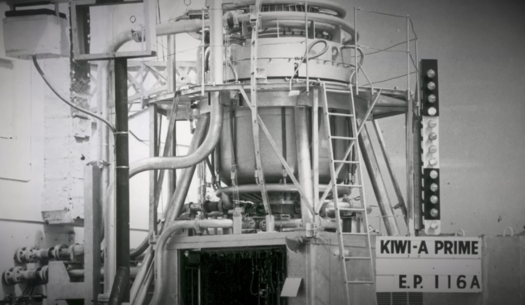 Black and white image of a complex machinery setup labeled "KIWI-A PRIME E.P. 116A." It features a large central chamber, pipes, and ladders, suggesting an industrial or research setting.