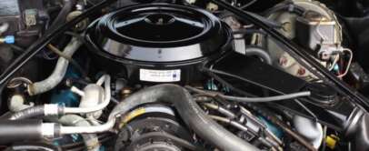 Close-up of a car engine showing various components including hoses, wires, and a round air filter housing at the top. The engine is clean and well-organized with a label visible on the air filter cover.