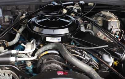 Close-up of a car engine showing various components including hoses, wires, and a round air filter housing at the top. The engine is clean and well-organized with a label visible on the air filter cover.