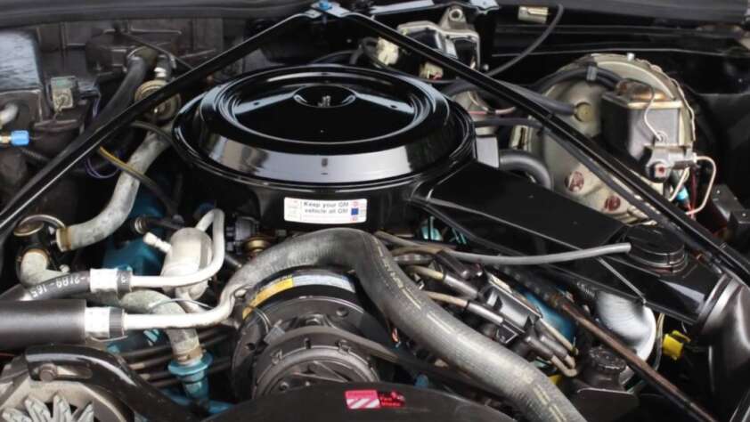 Close-up of a car engine showing various components including hoses, wires, and a round air filter housing at the top. The engine is clean and well-organized with a label visible on the air filter cover.