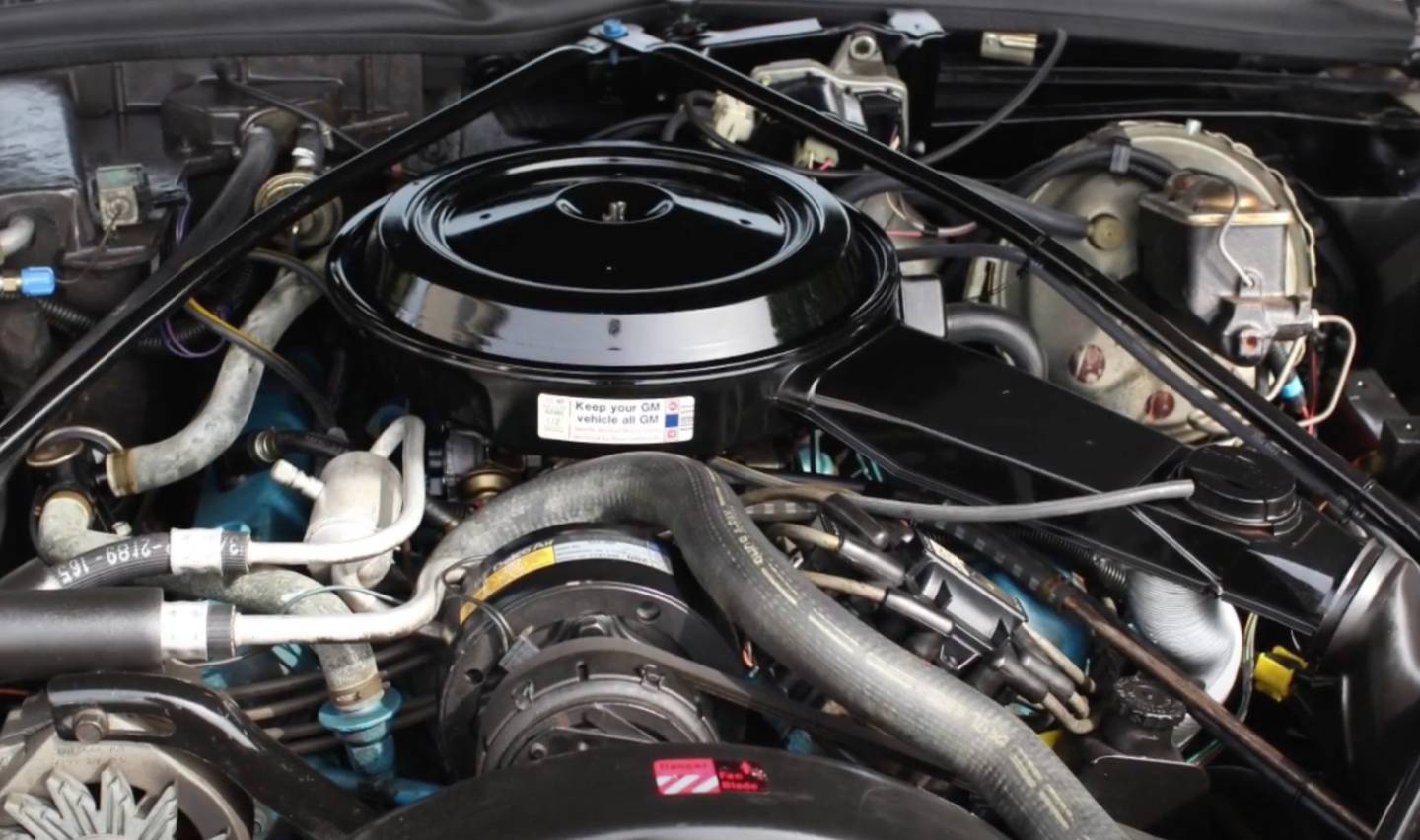 Close-up of a car engine showing various components including hoses, wires, and a round air filter housing at the top. The engine is clean and well-organized with a label visible on the air filter cover.