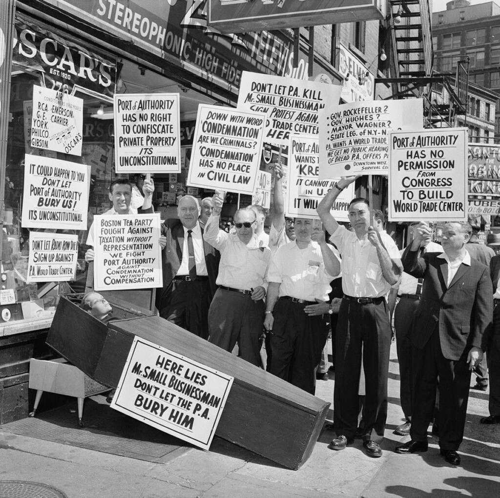 Black and white photo of a group of men holding protest signs against the Port Authority outside a store. A coffin on the sidewalk reads, "Here Lies Mr. Small Businessman." The protest emphasizes property rights and fair compensation.