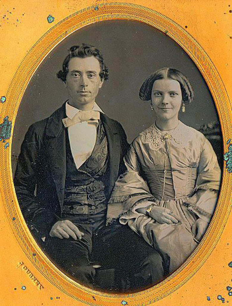 A vintage portrait of a man and woman seated side by side, dressed in 19th-century formal attire. The man wears a dark suit with a bow tie, and the woman wears a long dress with a lace collar. The image is set in an ornate oval frame.