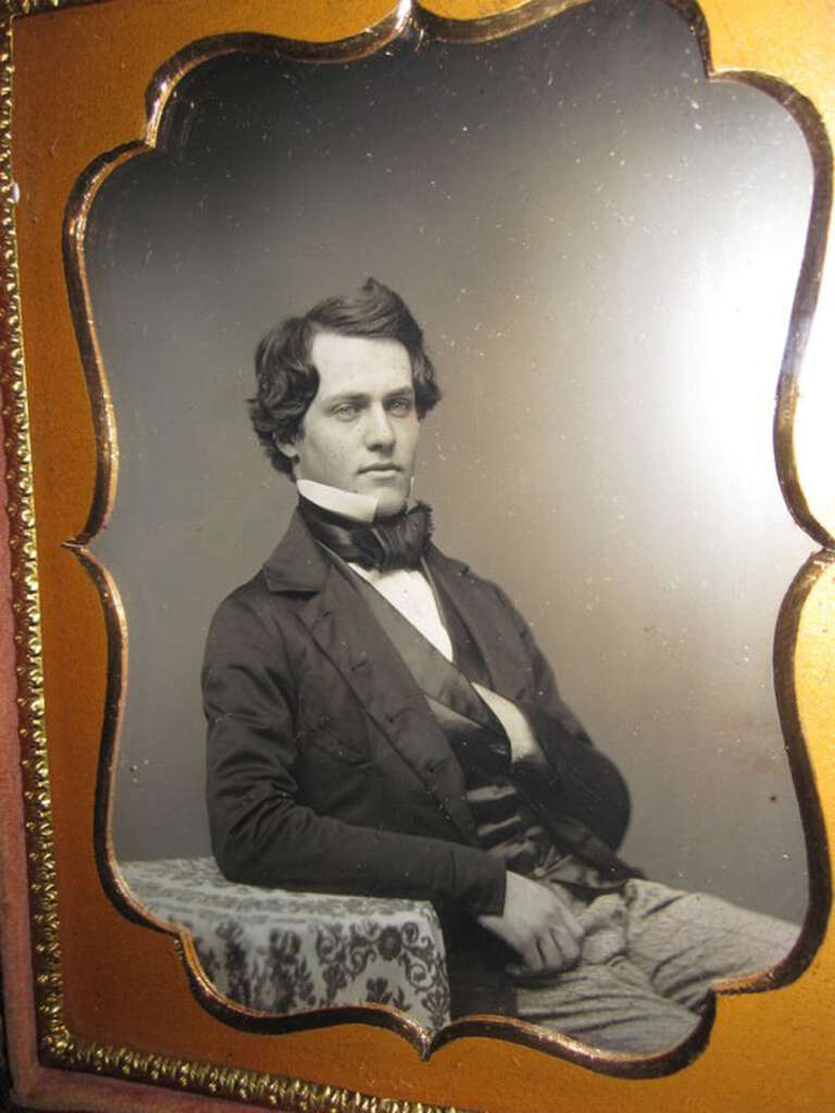 A sepia-toned portrait of a man in 19th-century attire. He has dark hair, a bow tie, and a formal jacket. His right hand is partially tucked into his waistcoat. The image is framed in an ornate, gold-edged border.