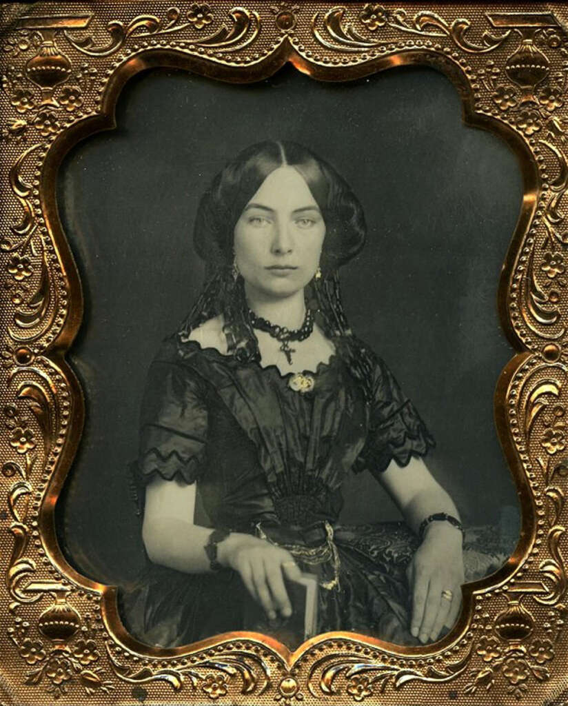 A woman in a black Victorian-era dress and intricate jewelry poses for a portrait. She has long, dark, braided hair and holds a small book in her left hand. The photo is framed in an ornate, gold-colored border with floral patterns.