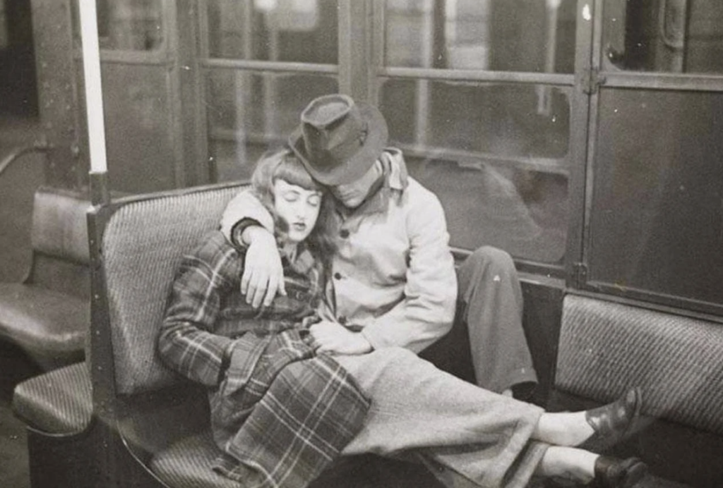 A black and white photo of a couple sitting on a subway bench. The woman is wearing a plaid coat, resting her head on the man's shoulder. The man in a hat and coat has his arm around her, both appearing relaxed and content.