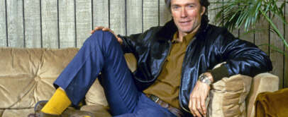 Clint Eastwood sits casually on a beige couch, wearing a black leather jacket, brown shirt, blue jeans, yellow socks, and brown shoes. The wooden wall and a green plant are in the background.